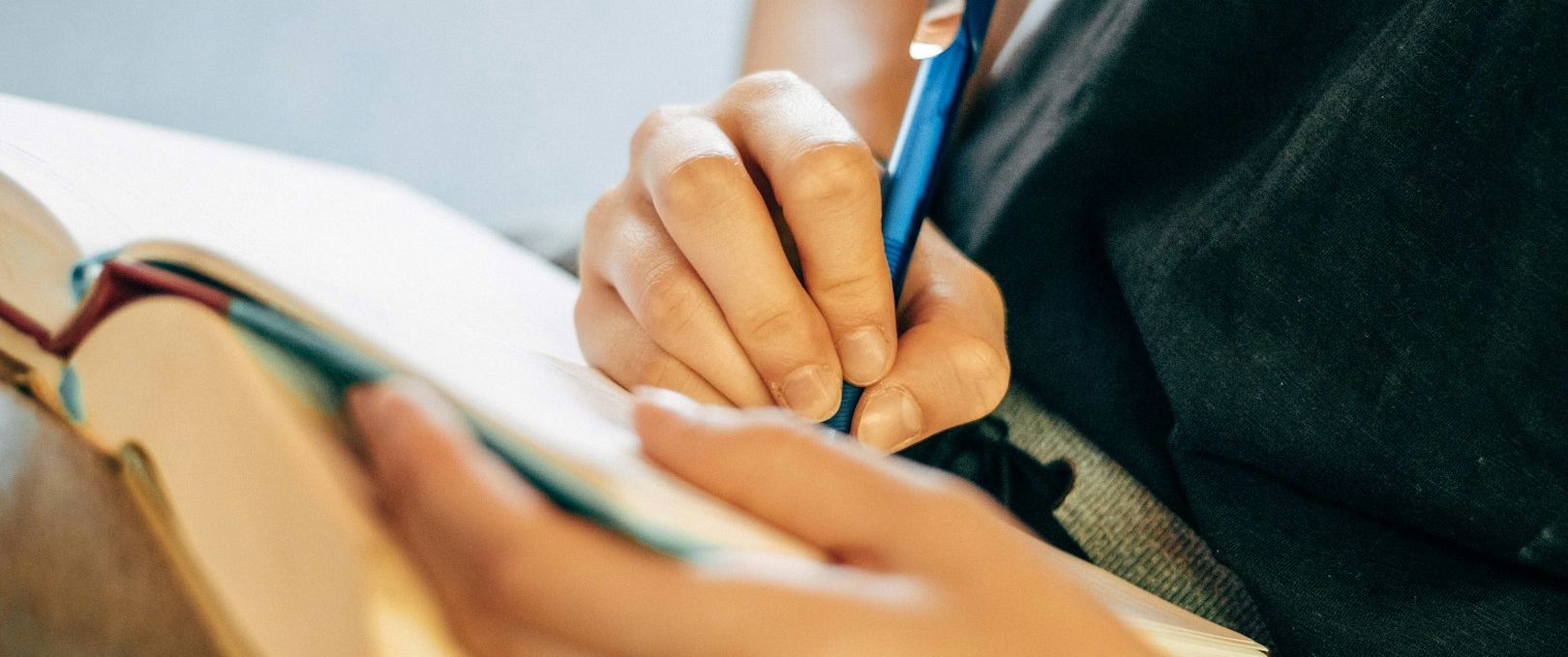 Auf dem Bild sieht man zwei Hände und ein offenes Heft. Die eine Hand hält einen Stift und beginnt, etwas in das Heft zu schreiben.